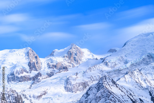 Snow-covered summits of Mont Maudit rise above rugged alpine ridges, illuminated by soft daylight under a vibrant blue sky with gentle cloud streaks.