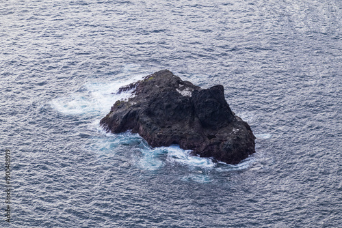 Isolated dark volcanic sea rock surrounded by vast ocean water and white foam waves.