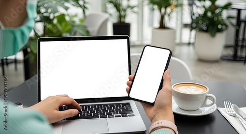 Person holding a smartphone and typing on a laptop in a cafe with plants and coffee