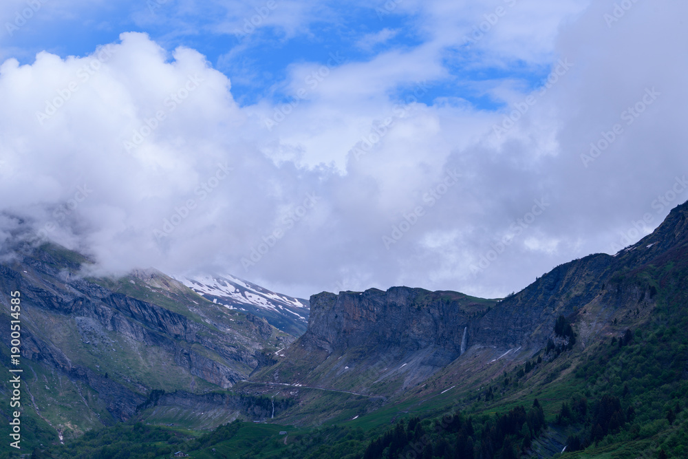 Fototapeta premium Steep rock faces rise above a lush green valley as low clouds drift overhead, with hints of snow and a waterfall visible in the dramatic mountain landscape.