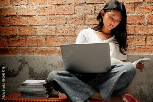 Southeast Asian young woman sitting cross-legged studying with a laptop and books, against a rustic brick wall background