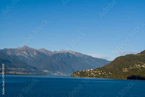 Quadro su tela Wide view of Cannobio perched on green hills above Lake Maggiore in northern Italy, with deep blue water and rugged alpine peaks under a vivid, cloudless sky