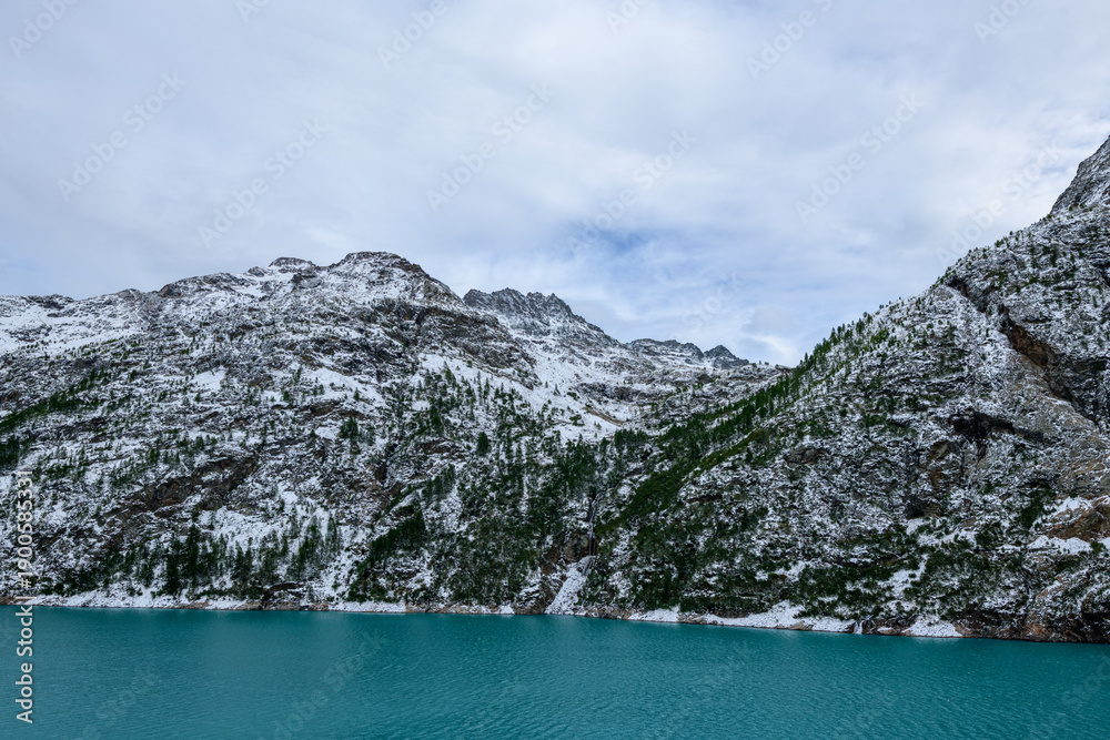 Obraz premium A vivid turquoise lake stretches beneath rugged, snow-dusted mountain slopes and scattered evergreen trees near Place-Moulin in the Aosta Valley, Italy. The dramatic alpine landscape is defined by