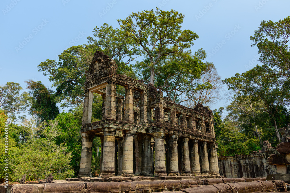 Obraz premium Two-story sandstone gallery with rows of columns stands amid dense tropical trees at Preah Khan temple in Cambodia's Angkor region. Weathered stone, mossy surfaces, and open blue sky evoke the