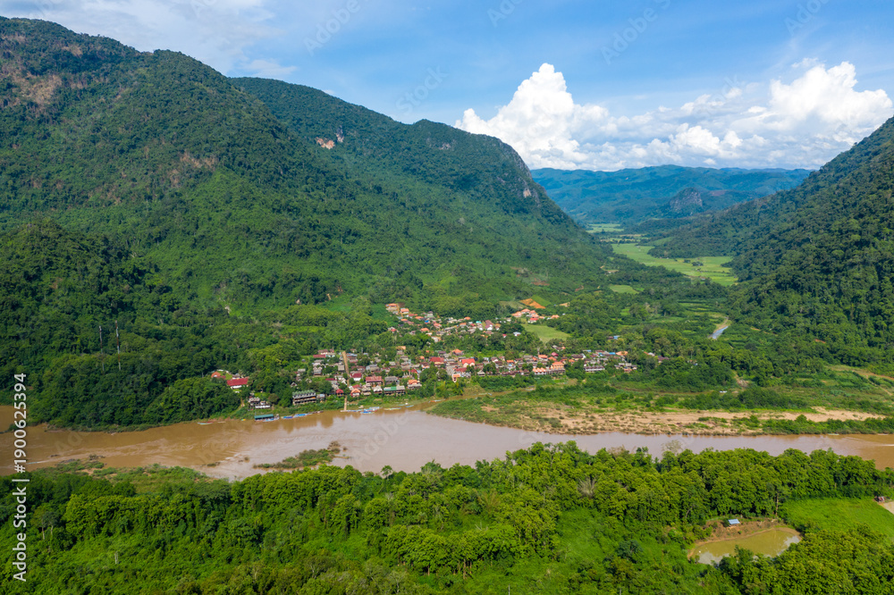 Fototapeta premium Aerial panorama of Muang Ngoi village nestled between steep green mountains and the muddy Nam Ou river in northern Laos. The scene features vibrant tropical vegetation, a broad river, and a dramatic