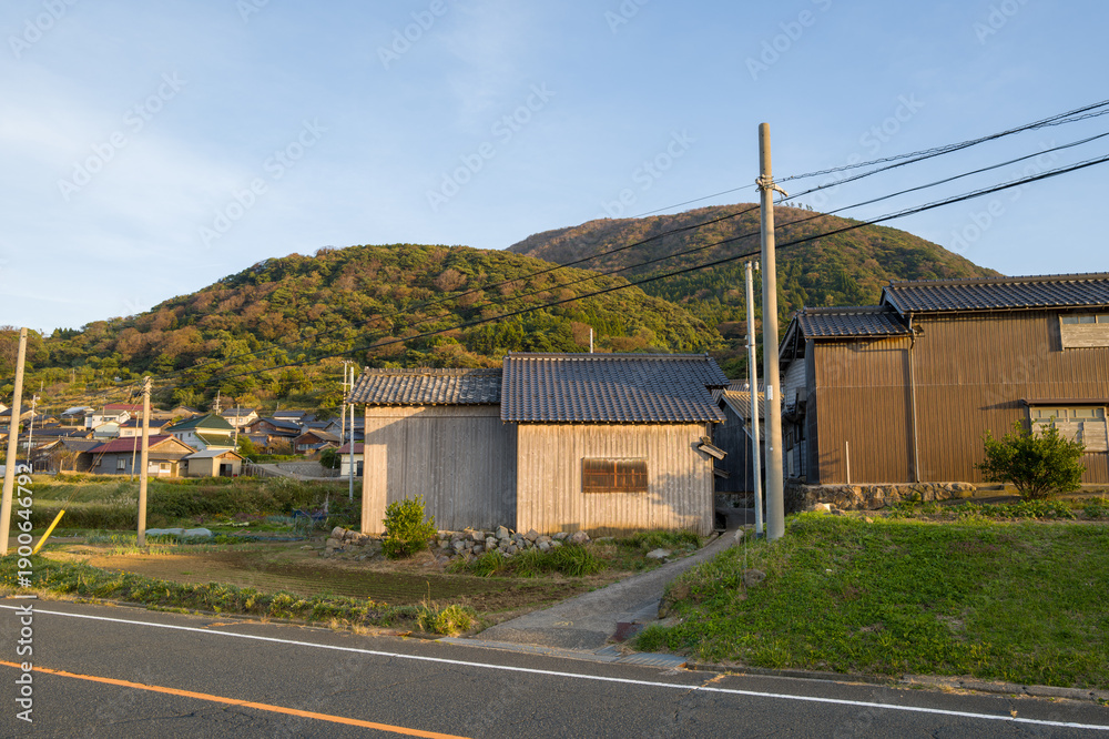 Obraz premium Wooden rural homes stand along a quiet roadside, backed by lush green hills under warm evening sunlight. The scene captures a peaceful village atmosphere with natural textures and soft shadows.
