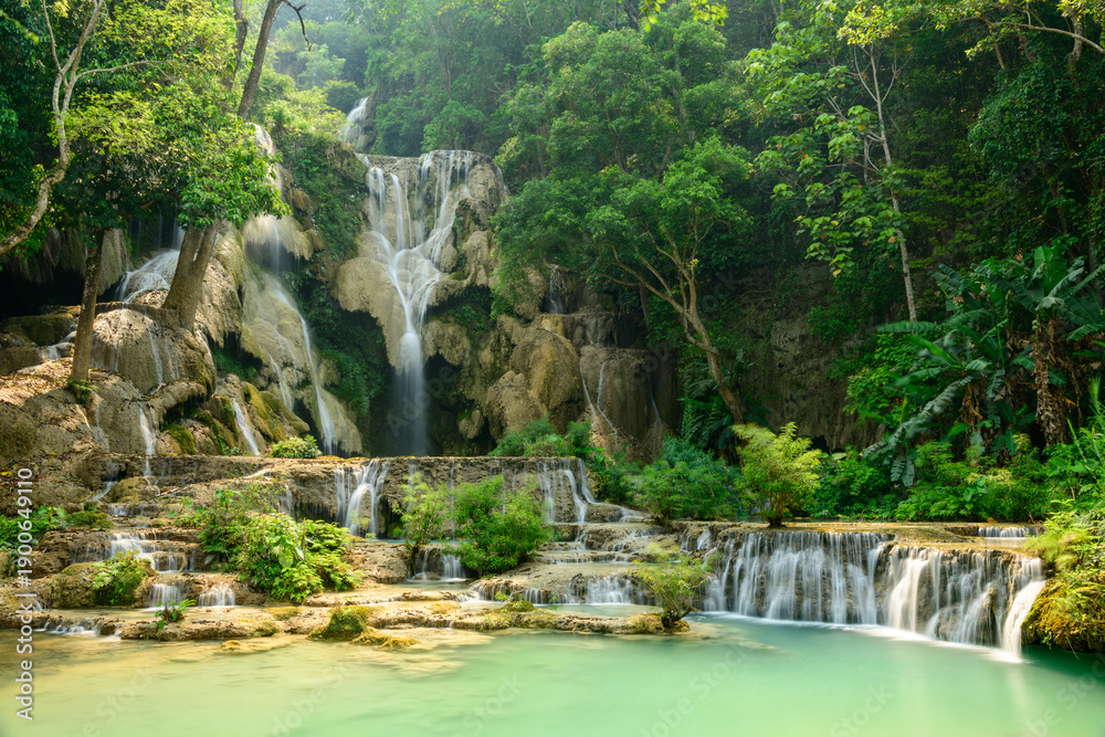 Naklejka premium Soft sunlight illuminates tiered limestone cascades and turquoise pools surrounded by dense tropical forest at Kuang Si Waterfalls in northern Laos. Lush greenery and flowing water create a tranquil
