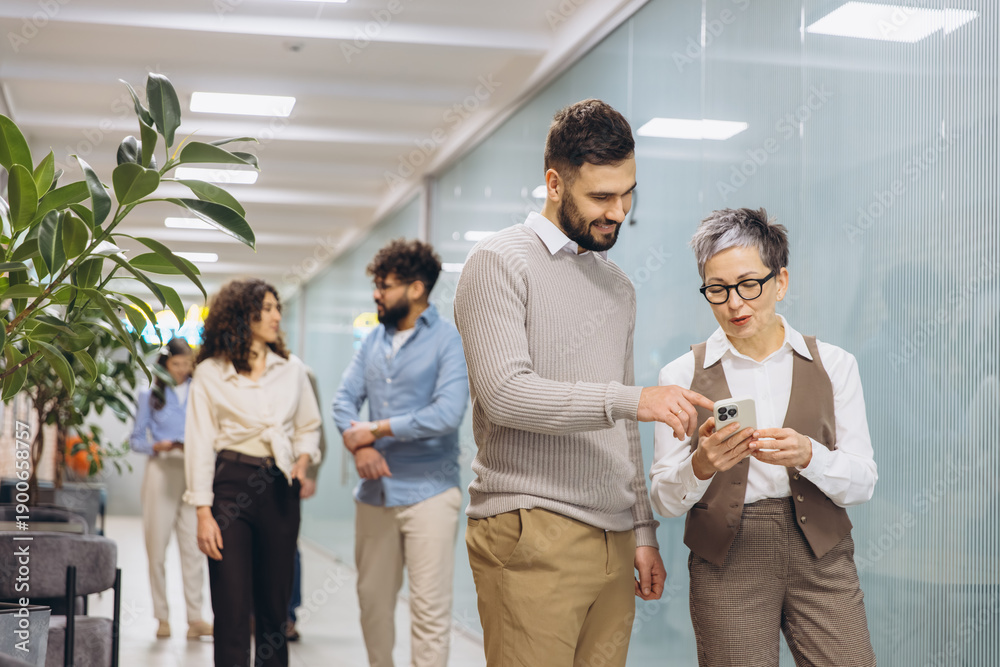 Obraz premium Diverse colleagues looking at smartphone, collaborating and discussing business strategy in an office hallway