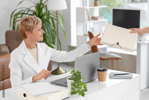 Beautiful mature businesswoman taking folder from secretary at table in office