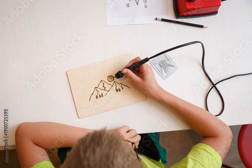 A child is using a pyrography pen to burn a mountain design onto a wooden board. Pencil, paper with sketches and a red pyrography tool are visible on the table - a creative craft session