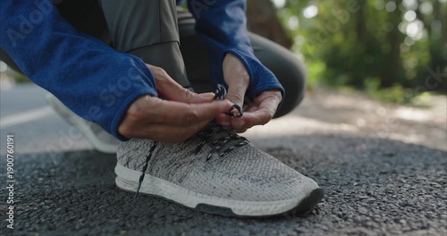 Ground level view shows a woman kneeling, tightening black and white laces on a light gray SPORT running shoe in a public park with golden evening light and bokeh.