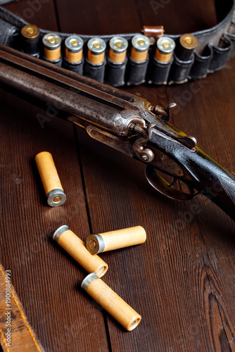 Vintage shotgun resting on a wooden surface, surrounded by scattered shotgun shells and a leather ammunition belt, showcasing the craftsmanship and history of firearms