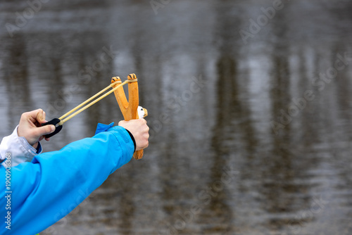 Fotografie Person holding wooden slingshot with elastic bands, aiming toward water surface