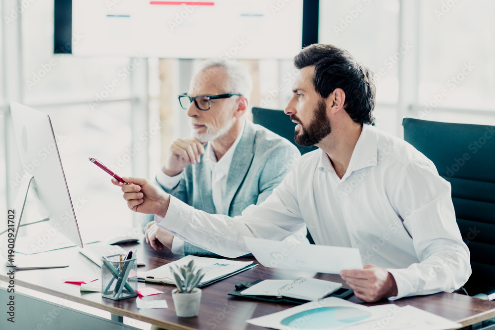© deagreez - Business meeting in modern office two colleagues discuss documents and strategy at a sleek desk while one man points at a computer screen for a productive corporate discussion