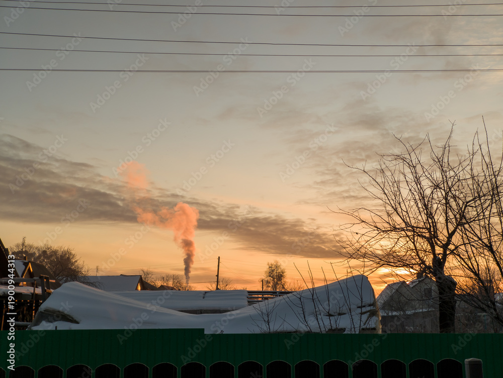 Obraz premium Winter Sunset: Snowy Village with Smoke Plume and Sunset Light