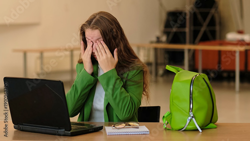 A young student pauses and covers her eyes at a desk with a laptop and notebook, side angle framing and calm tones. It relates to homework deadlines, fatigue, and coping with academic stress.