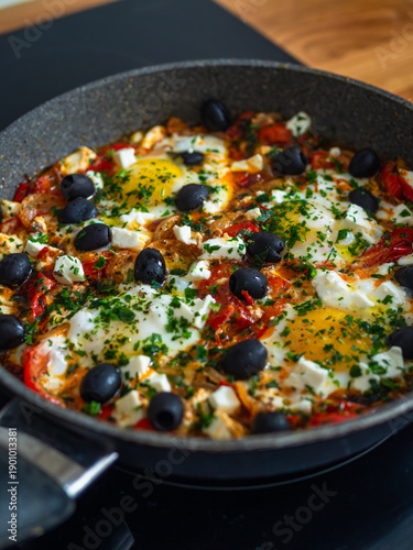Shakshuka - fried eggs with vegetables and black olives in frying pan on electric stove