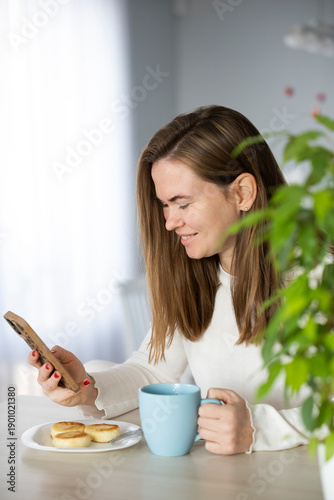 Young woman in white pajama with mobile phone and breakfast at home in modern morning routine. Using smartphone, checking messages, working from home, reading news. Digital lifestyle.