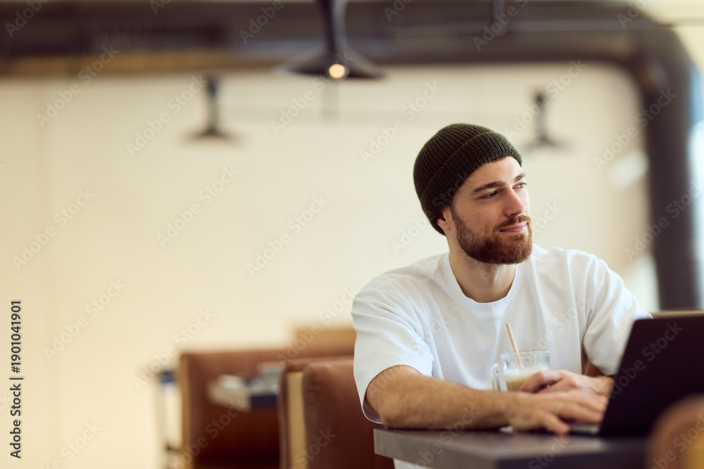 Obraz premium Casual Diner Workspace: Bearded Man in White Tshirt and Beanie Working on Laptop