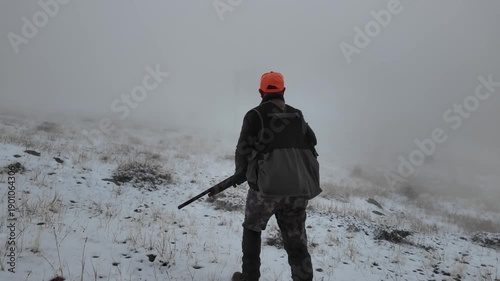 Rear view of a hunter carrying a rifle while walking through a foggy, snowy natural landscape. Represents hunting, wilderness, rural life, outdoor activity, and survival atmosphere.