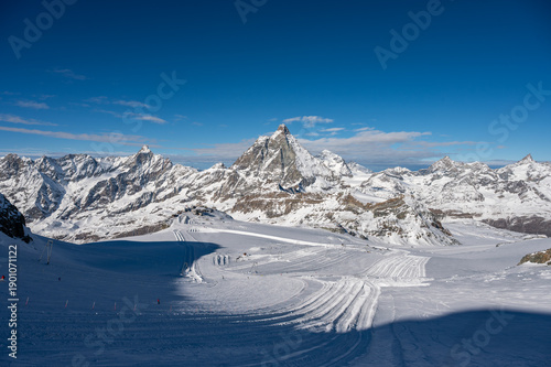 Daytime view of the Plateau Rosa ski area at Testa Grigia, featuring skiers, snow-covered alpine terrain, and the Matterhorn under a clear blue sky.