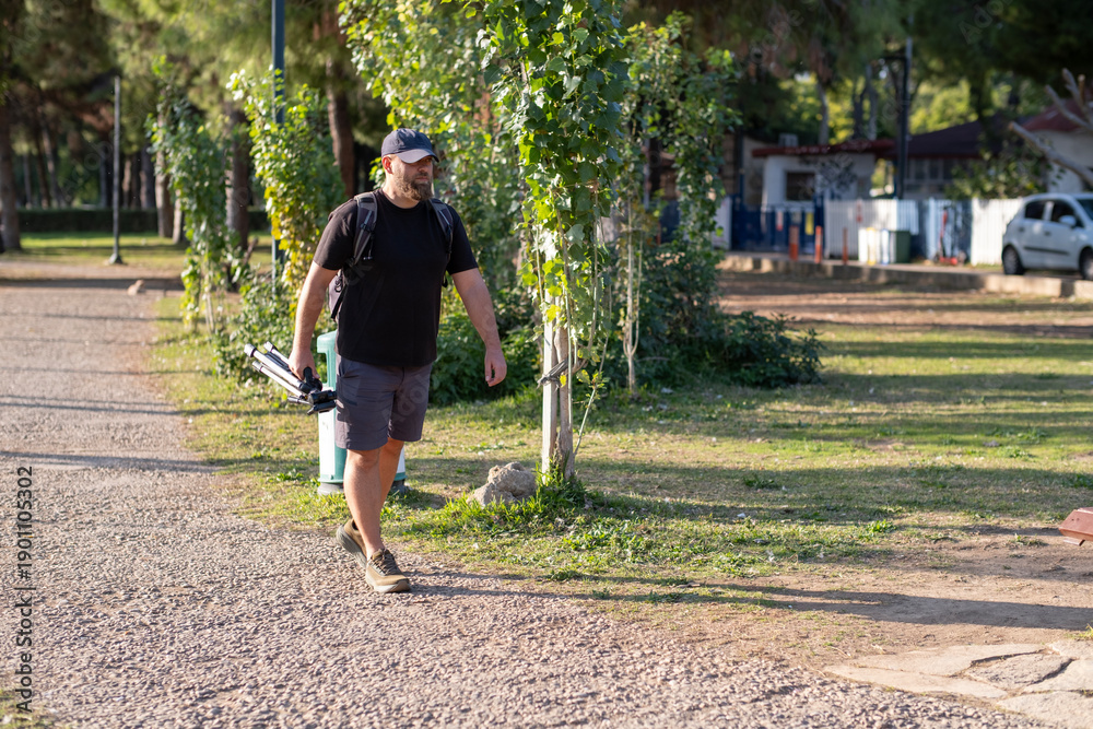 Fototapeta premium Photographer Walking Through Park Carrying a Tripod. Male Photographer Holding Tripod While Walking in Park. Man Carrying Tripod During Outdoor Photo Session.