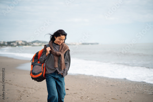 Fotografía Smiling woman walking along sandy beach with orange backpack, casual travel life