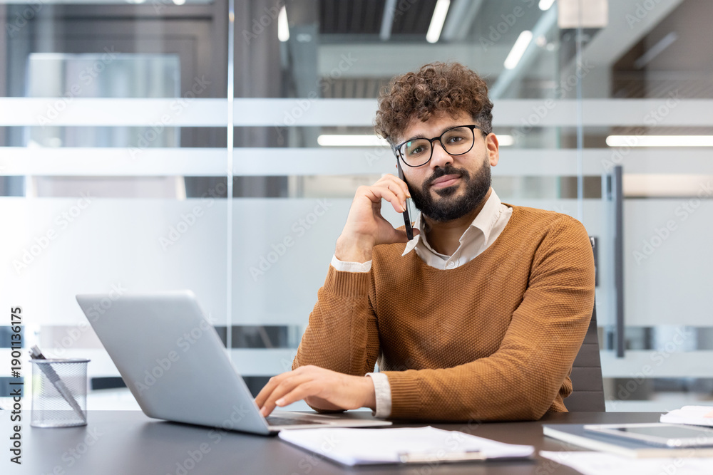 Fototapeta premium Young businessman with curly hair and eyeglasses communicating on his mobile phone while working on a laptop at a contemporary workspace, managing business operations