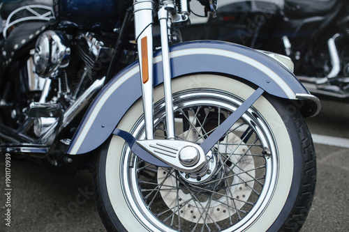 Obraz na plátně Detailed shot of a vintage-style motorcycle front end, featuring a metallic blue fender with white pinstripes, a classic whitewall tire, polished chrome wire-spoke wheels, and chrome front forks