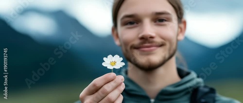 Man holds small white flower in outdoor setting near mountains during sunny weather