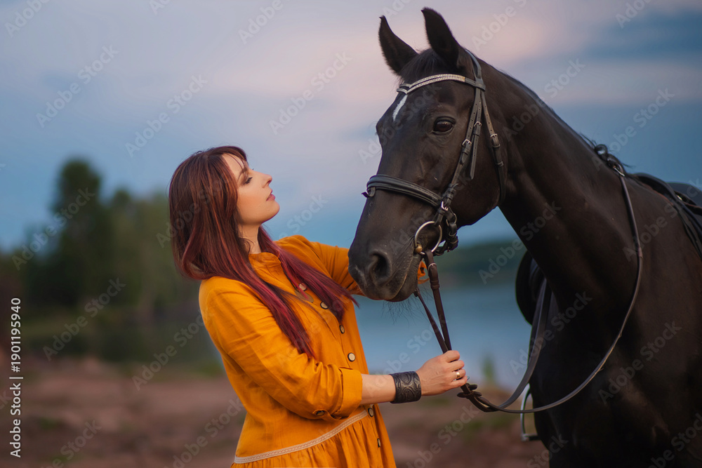 Obraz premium A woman with beautiful hair in an orange dress tenderly hugs a horse against the backdrop of a calm sky and a lake. Photo shoot by the water.