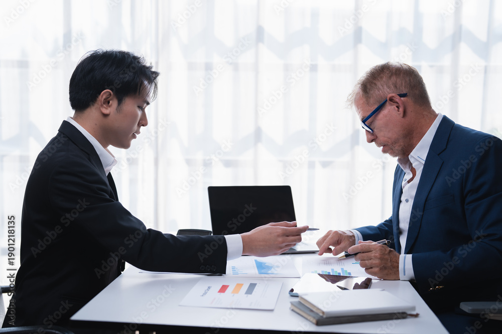 Fototapeta premium Two business men in suits reviewing financial reports and discussing strategy during a corporate meeting in a modern office setup, collaborating on analytics and planning