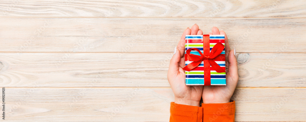 Fototapeta premium Female's hands holding striped gift box with colored ribbon on yellow rustic wooden background. Christmas concept or other holiday handmade present box, concept top view with copy space