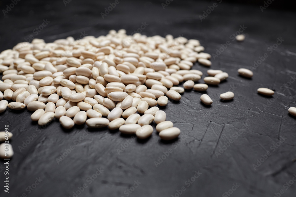 Fototapeta premium White beans scattered on a dark textured surface, showcasing their smooth, oval shapes and natural color variations in a close-up view