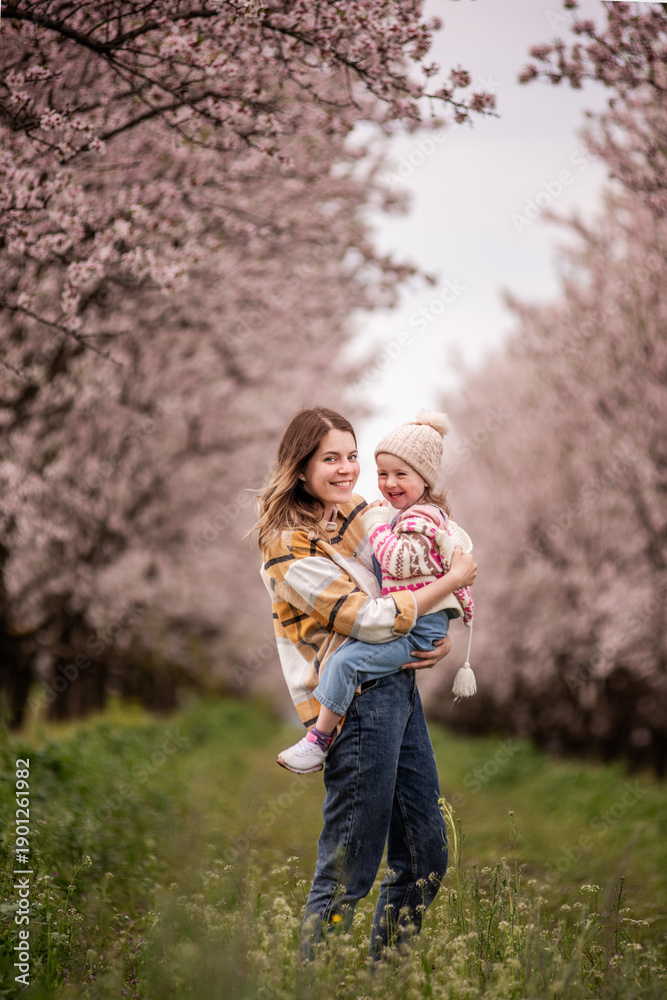 Fototapeta premium Mother joyfully holding her laughing daughter in a sunlit meadow of green grass and pink blossoms, sharing a tender springtime moment of love, laughter and carefree bonding