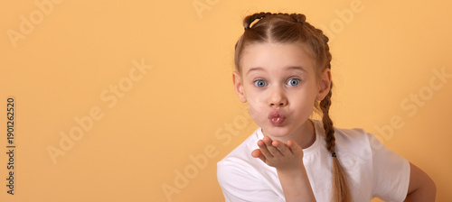 Schilderij op canvas Adorable young girl in white t-shirt blowing a kiss with playful expression, isolated on pastel orange background, copy space