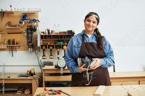 Canvas Print Smiling young woman stands confidently in a workshop holding tools, ready to cre