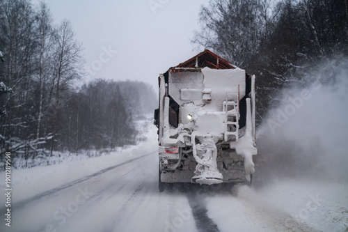 A heavy utility truck clears a snowy road by spreading salt and sand creating a white cloud of dust during a cold winter day on a highway