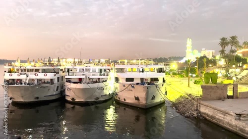 Tour boats moored at a river harbor during nighttime with city lights reflected on the water. Urban waterfront scene with illuminated buildings and boats.