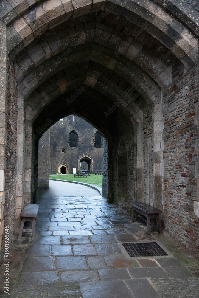 Obraz premium A wide-angle interior view looking through a massive stone vaulted archway of a medieval castle, leading to a cobblestone courtyard with wooden benches and historic ruins in the background.