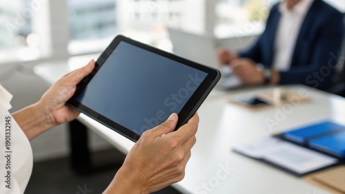 A person holds a tablet in a modern office, with a colleague visible in the background, illustrating technology in a business environment.