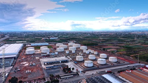 Approaching a large territory of a modern oil refinery plant with diverse size tanks for fuel storage. Aerial view. Green fields and vast cityscape at backdrop.