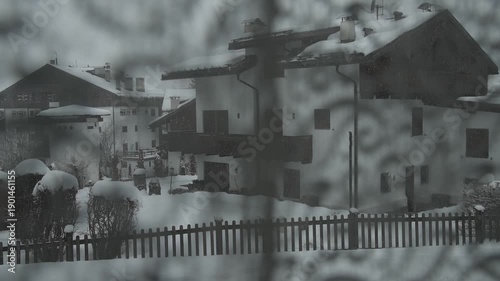 Viseo of snow-covered alpine houses sit quietly during a winter storm, viewed through window with curtains. Soft snowfall, muted light, and wooden chalets create a calm, cold, and secluded mountain