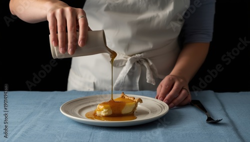 Anonymous woman drizzling caramel over cheese pie on a blue table, set against a dark backdrop after making the dessert at home