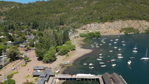 Aerial view of Lake Lacar and San Martin de los Andes in Patagonia, highlighting a scenic marina with numerous sailboats and boats anchored in deep blue water surrounded by mountains.