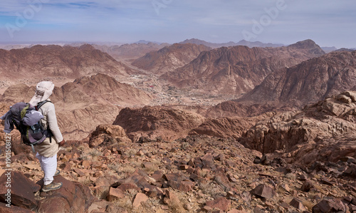 A bedouin in traditional clothing, long white robe with headscarf, in a view point to the town Saint Catherine in Sinai. Panorama of high red mountains around the town. Authentic bedouin lifestyle.