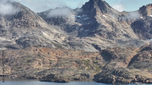 Cinematic aerial shot of steep, rocky mountains in the Arctic circle. High-altitude drone view showing the raw beauty of East Greenland's geology, with mist clinging to the summits under a blue sky.