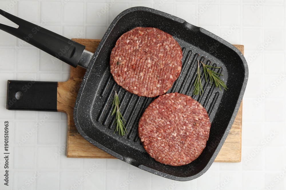 Fototapeta premium Uncooked meat patty with rosemary in frying pan on white tiled table, top view