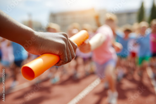 Close-up of child passing bright orange baton during a relay race on outdoor track with other children running in blurred background on sunny day