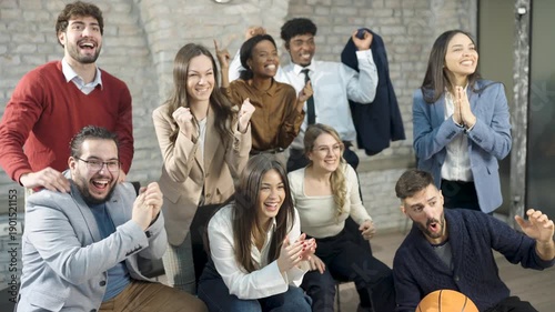 Medium group of young, diverse office colleagues watching a basketball game indoors. One person holds a basketball while everyone cheers, capturing fun and teamwork in a casual workplace.